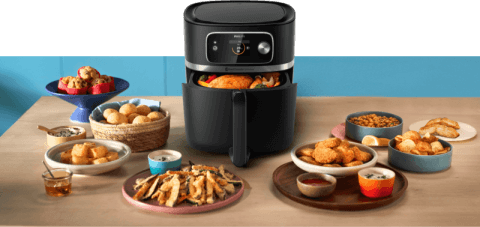 Na stole znajduje się czarna frytkownica beztłuszczowa z wyświetlaczem, wokół której rozmieszczone są różne potrawy, takie jak frytki, kurczak i pieczywo. W tle widoczne są kolorowe miski z jedzeniem.