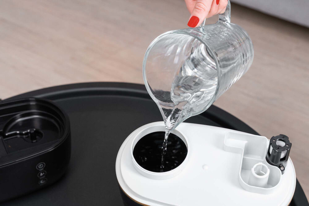 Nawilżacz GÖTZE & JENSEN AH250K Powiększone ujęcie dłoni z czerwonym lakierem na paznokciach, nalewającej wodę z przezroczystego dzbanka do zbiornika nawilżacza. Widoczny jest biały element wewnątrz czarno-złotej obudowy.