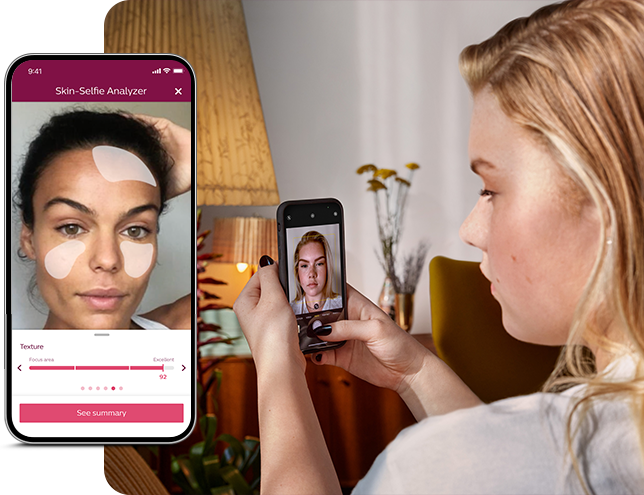 Osoba trzyma smartfon z aplikacją \'Skin-Selfie Analyzer\', która pokazuje twarz z nałożonymi filtrami. W tle widać lampę i rośliny na stole.