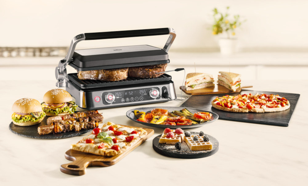 Na blacie kuchennym znajduje się grill elektryczny z otwartą pokrywą, w którym grillowane są kawałki mięsa. Wokół grilla rozmieszczone są różnorodne potrawy, takie jak kanapki, pizza, gofry z owocami i grillowane warzywa.