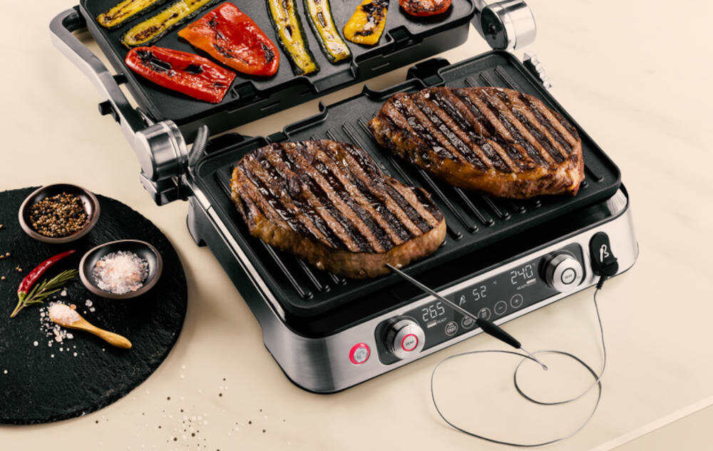 Grill elektryczny z otwartą pokrywą, na której znajdują się grillowane warzywa, a na dolnej płycie dwa kawałki mięsa z termosondą. Obok grilla leży okrągła deska z przyprawami i papryczkami.