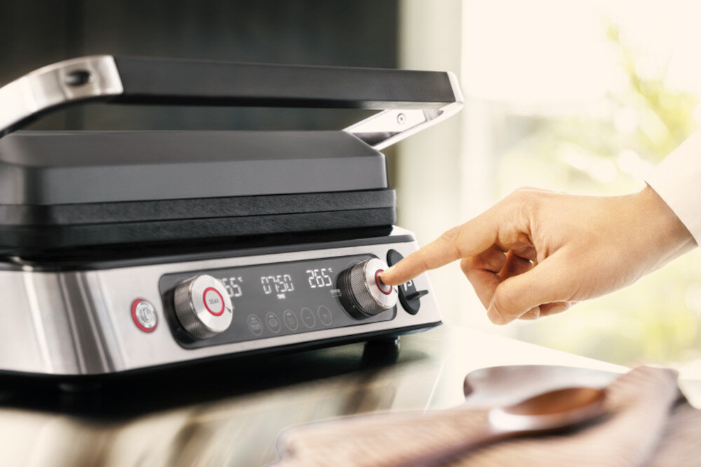 Osoba naciska przycisk na panelu sterowania elektrycznego grilla z wyświetlaczem cyfrowym pokazującym temperaturę 265°C. Grill ma metaliczną obudowę i czarne elementy.