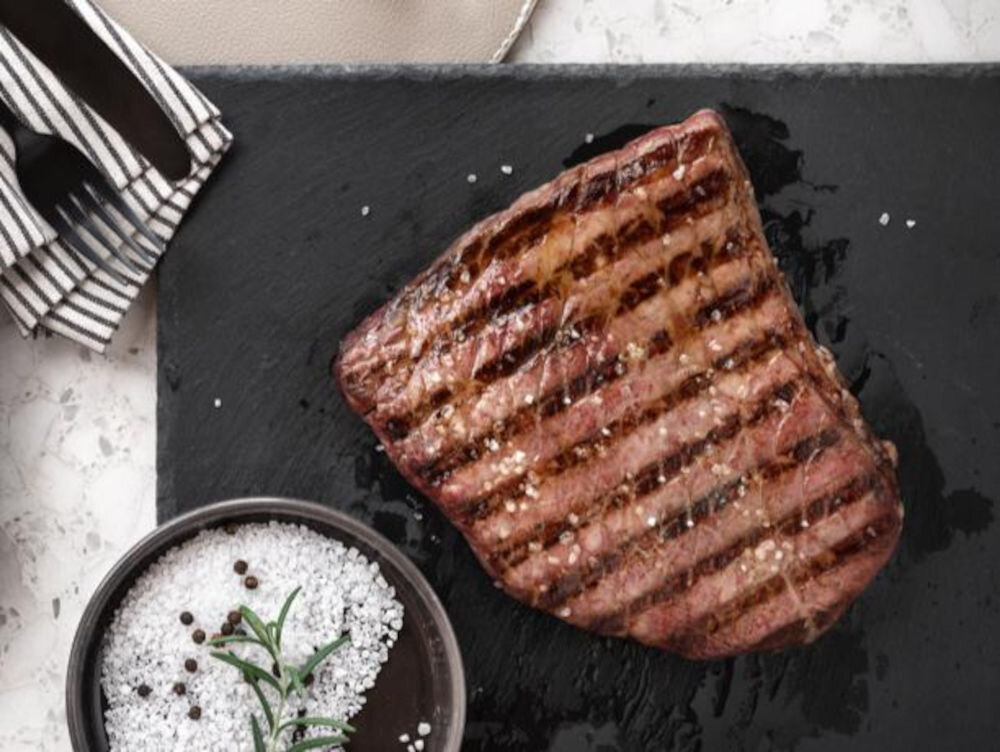 Na czarnej desce leży grillowany kawałek mięsa z widocznymi śladami rusztu. Obok znajduje się miseczka z solą i gałązką rozmarynu.