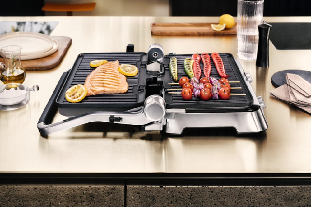 Na blacie kuchennym znajduje się otwarty grill elektryczny z grillowanym łososiem i warzywami na szpikulcach. Obok grilla leżą akcesoria kuchenne, w tym szklanka z wodą i przyprawy.