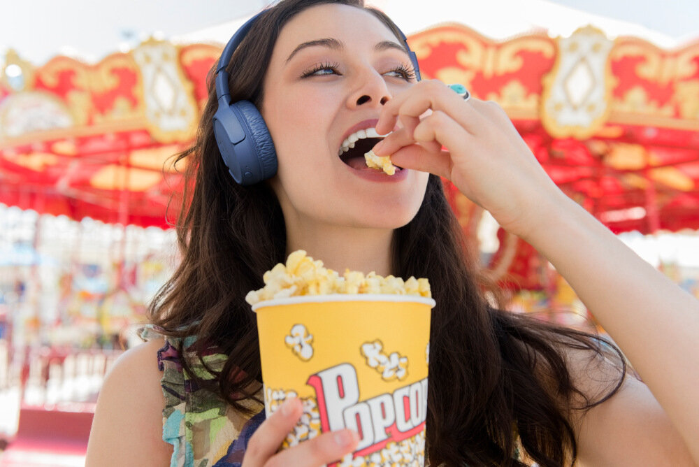 Osoba nosi niebieskie słuchawki nauszne i je popcorn z żółtego kubka z napisem \'Popcorn\'. W tle widoczna jest kolorowa karuzela.