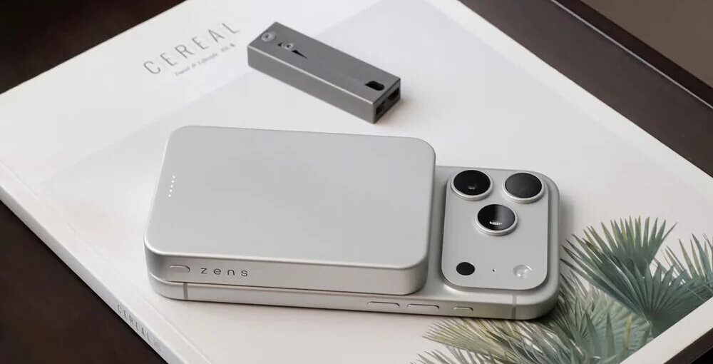 Na białej powierzchni leży srebrny powerbank z logo \'zens\' oraz smartfon z trzema obiektywami aparatu. W tle widoczna jest książka z napisem \'CEREAL\'.