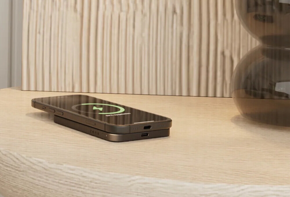 Na drewnianym blacie leży czarny powerbank z telefonem na wierzchu, na ekranie telefonu widoczna jest ikona ładowania. W tle znajduje się dekoracyjny element z ciemnego szkła.
