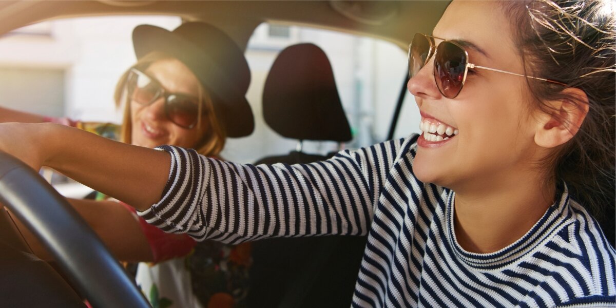 Dwie osoby siedzą w samochodzie, obie uśmiechnięte i noszące okulary przeciwsłoneczne. Osoba z przodu ma na sobie bluzkę w paski.