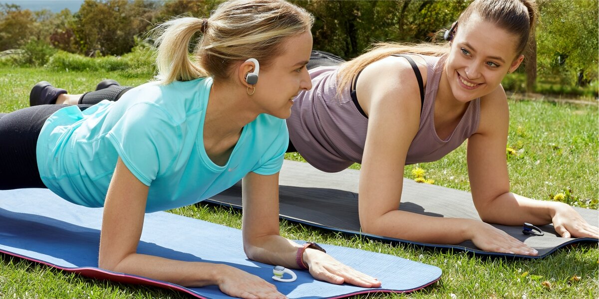 Energia w rytmie muzyki Dwie kobiety wykonujące plank (ćwiczenie deska) na matach do jogi na świeżym powietrzu. Jedna z nich ma w uchu biało-srebrną słuchawkę sportową, co sugeruje wygodę noszenia podczas ćwiczeń.