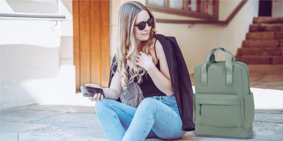 Plecak na laptopa AMERICAN TOURISTER Soulpack 15 cali  Iceberg Green Zielony Młoda kobieta w okularach przeciwsłonecznych siedząca na schodach obok szałwiowego plecaka American Tourister w miejskim otoczeniu.