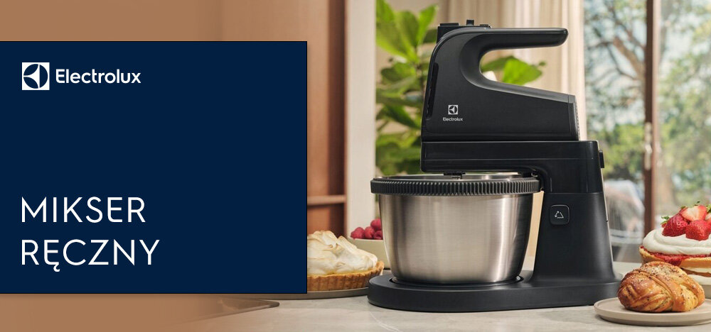 Na obrazie widoczny jest czarny mikser ręczny z metalową misą, stojący na blacie kuchennym. Obok znajdują się wypieki, a w tle widać roślinność i okno.