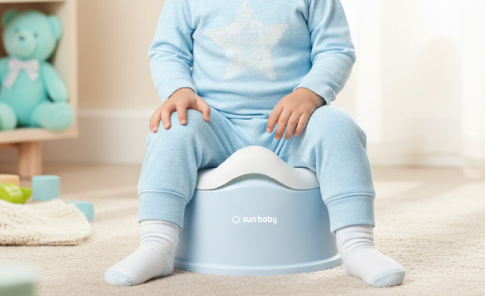 Dziecko w niebieskim ubraniu siedzi na biało-niebieskim nocniku z logo \'sun baby\'. W tle widoczne są zabawki i jasne zasłony.