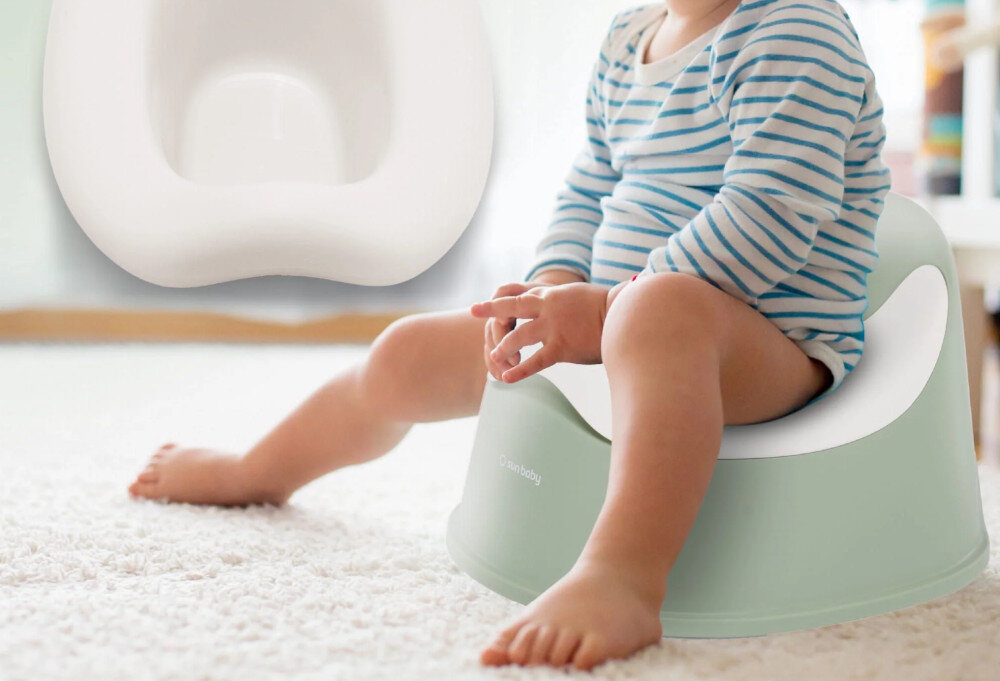Dziecko siedzi na biało-zielonym nocniku na jasnym dywanie. Na nocniku widoczne jest logo \'sun baby\'.