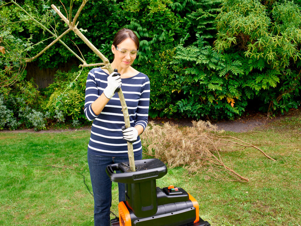 Rozdrabniacz do gałęzi BLACK&DECKER BEGAS5800-QS 2800W 40mm ogród trawnik osoba upycha gałęzie w rozdrabniaczu w tle drzewa krzewy przetwarzanie gałęzi do 45 mm średnicy mechanizm tnąco-rozdrabniający kompostowanie utylizacja