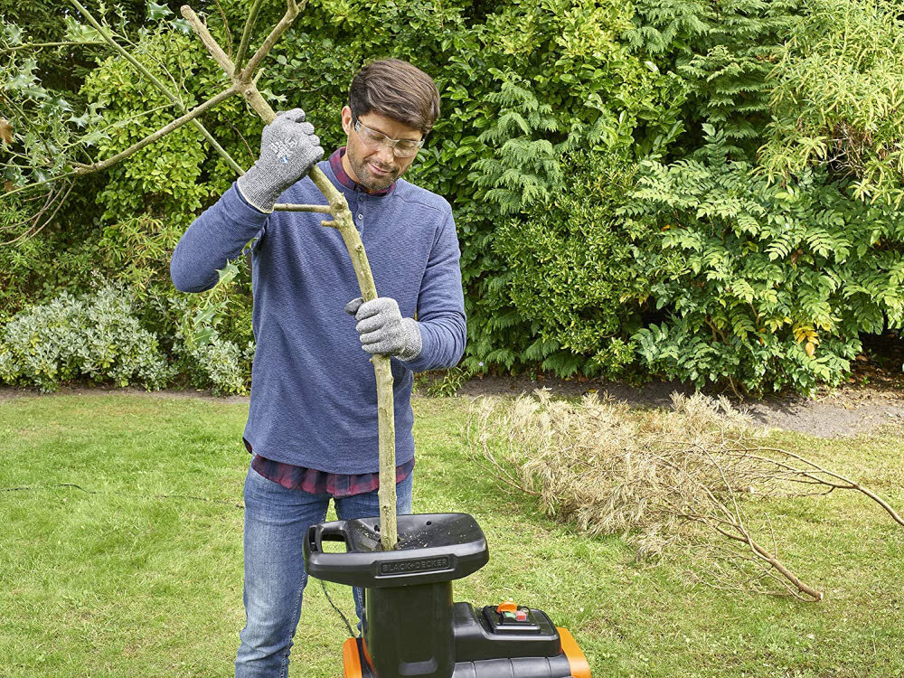 Rozdrabniacz do gałęzi BLACK&DECKER BEGAS5800-QS 2800W 40mm ogród trawnik osoba upycha gałęzie w rozdrabniaczu w tle drzewa krzewy mechanizm tnąco-rozdrabniający tnie i kruszy gałęzie usprawnia transport ogranicza konieczność ręcznego docinania gałęzi