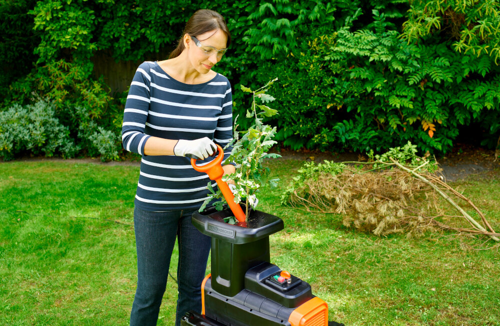 Rozdrabniacz do gałęzi BLACK&DECKER BEGAS5800-QS 2800W 40mm ogród trawnik osoba upycha w rozdrabniaczu gałęzie za pomocą popychacza w tle drzewa krzewy praktyczny popychacz bezpieczeństwo pracy ergonomiczna konstrukcja