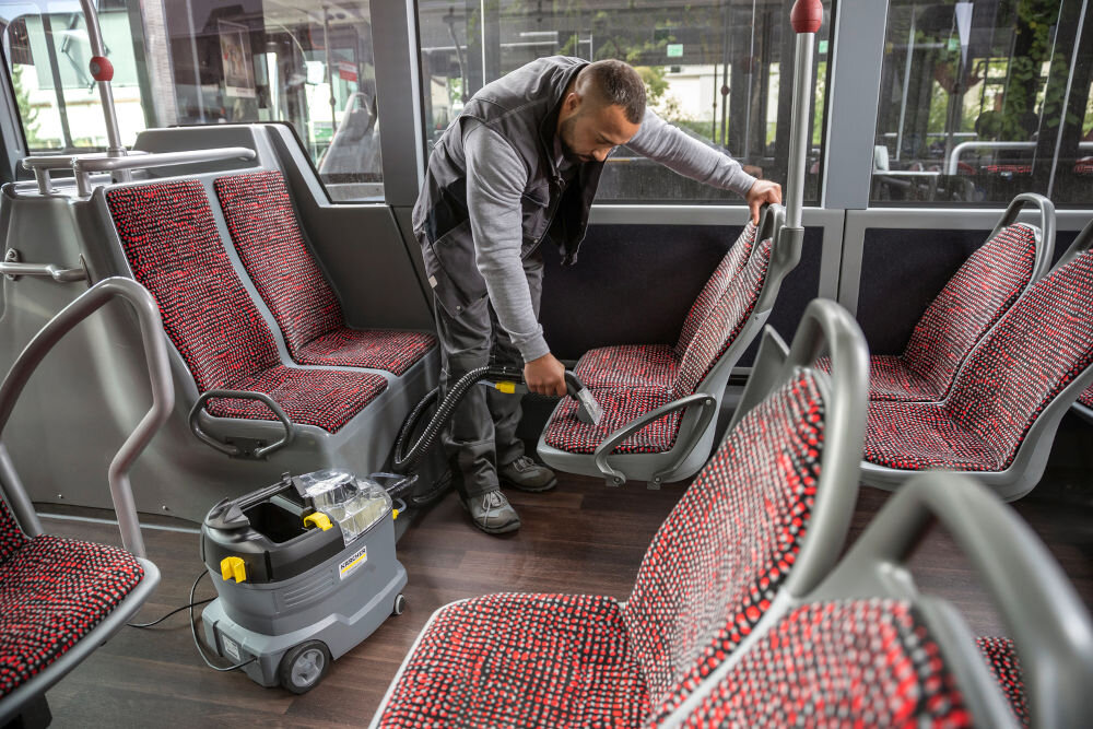 Osoba używa urządzenia ekstrakcyjnego do czyszczenia siedzeń w pojeździe komunikacji miejskiej. Obok znajduje się urządzenie z widocznym logo KARCHER.