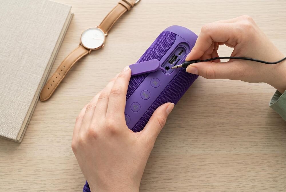 Osoba podłącza kabel do fioletowego głośnika mobilnego z widocznymi przyciskami i portami. Obok leży zegarek na brązowym pasku i zamknięty notes.