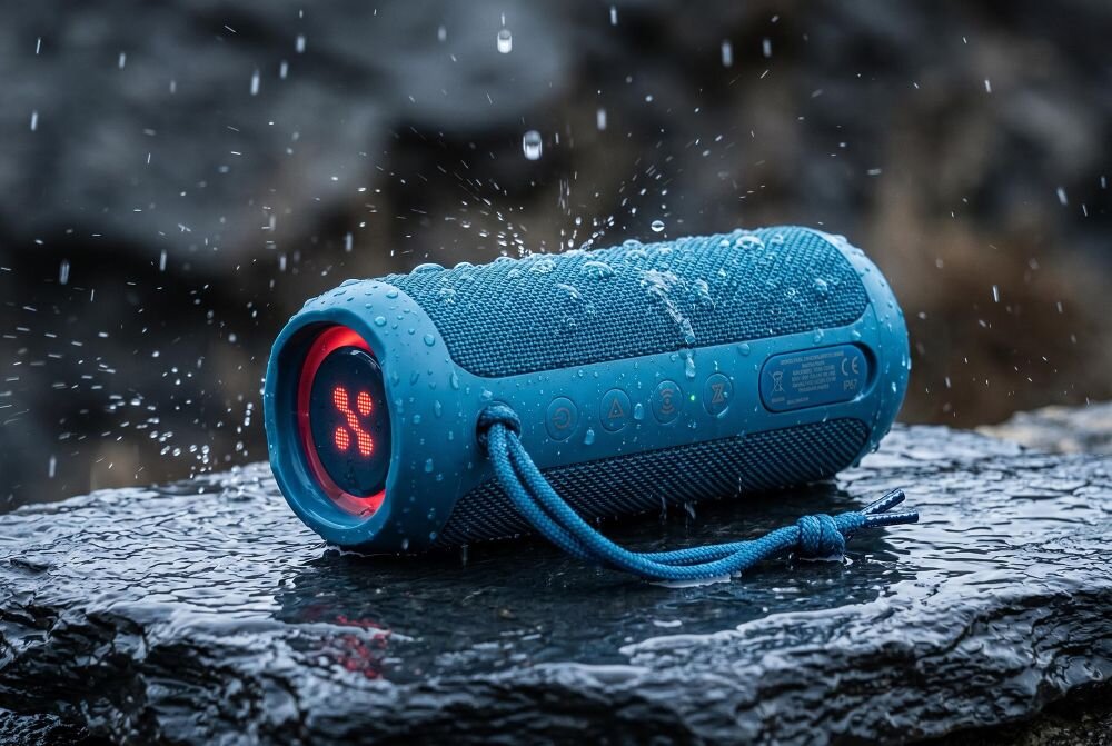 Niebieski głośnik mobilny leży na mokrej powierzchni, a na jego obudowie widać krople wody. Na jednym z końców głośnika świeci się czerwony symbol w kształcie litery X.