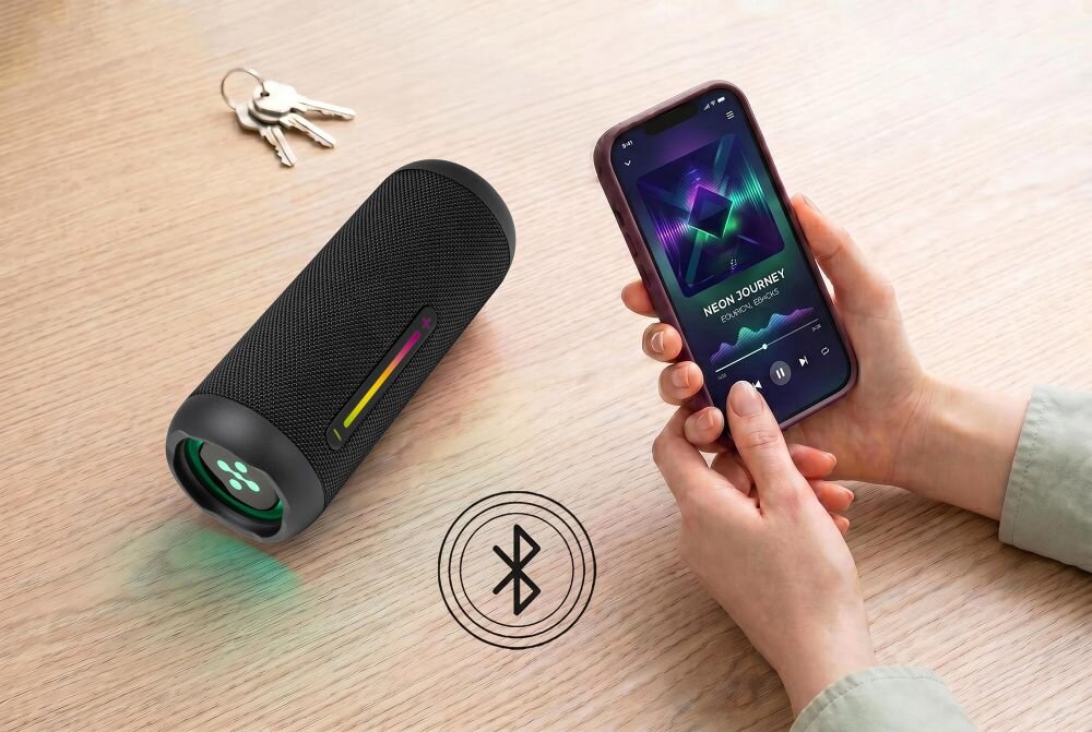 Na drewnianym stole leży czarny głośnik z kolorowym podświetleniem oraz smartfon z odtwarzaczem muzyki. Obok widoczne są klucze i symbol Bluetooth na powierzchni stołu.