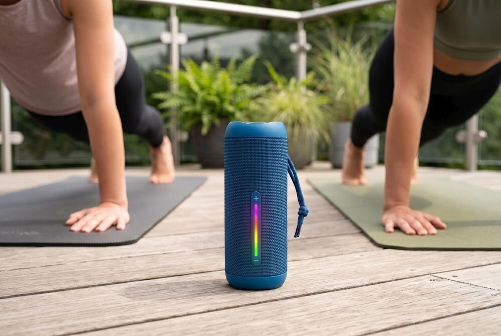 Na drewnianej podłodze stoi niebieski głośnik mobilny z podświetlanym panelem kontrolnym. W tle widać dwie osoby wykonujące ćwiczenia na matach oraz rośliny w doniczkach.