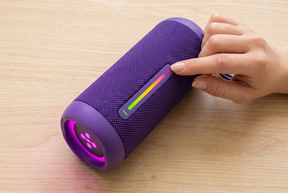 Fioletowy głośnik mobilny z podświetleniem LED, na którym widoczny jest panel dotykowy z kolorowym wskaźnikiem. Osoba dotyka panelu, a głośnik leży na drewnianej powierzchni.
