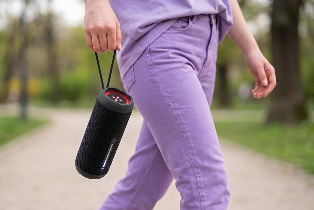 Osoba trzyma w ręce czarny, cylindryczny głośnik z czerwonymi elementami na górze. W tle widoczna jest ścieżka w parku.