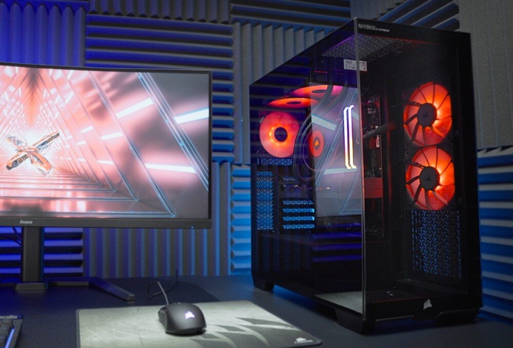 Komputer OPTIMUS E-Sport GB760T-CR17 Stanowisko gracza z włączonym komputerem stacjonarnym o przeszklonej obudowie, obok którego stoi monitor wyświetlający geometryczną grafikę, a na podkładce leży czarna mysz komputerowa, opis