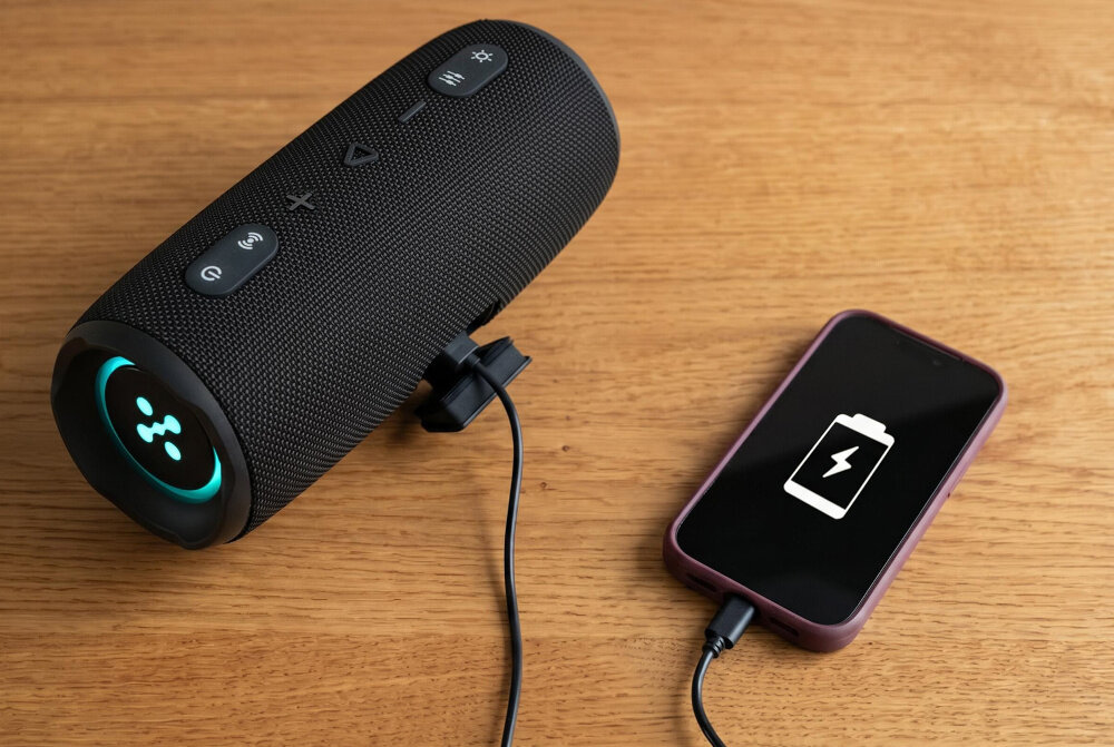 Na drewnianym blacie znajduje się czarny głośnik mobilny z kilkoma przyciskami oraz smartfon z ikoną ładowania baterii na ekranie. Oba urządzenia są połączone kablem USB.