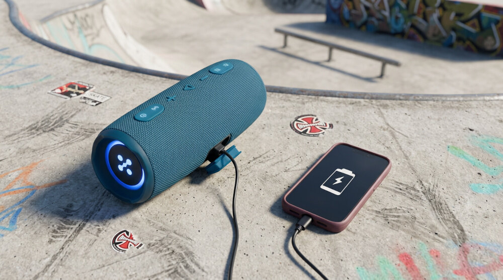Na betonowej powierzchni znajduje się niebieski głośnik mobilny z podświetlanym panelem oraz smartfon z ikoną baterii, oba połączone kablem. W tle widoczne są elementy skateparku i graffiti.