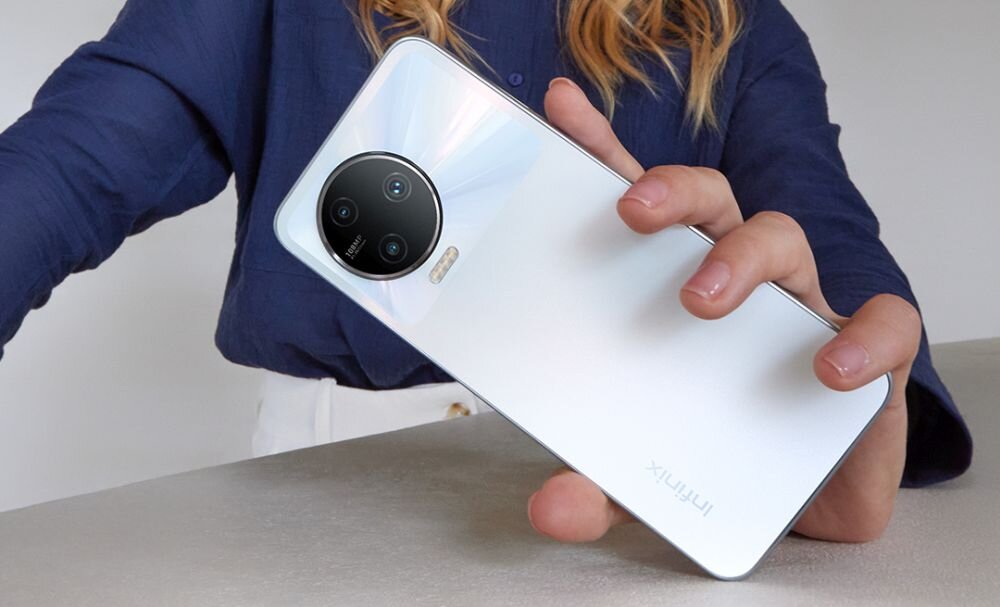 Osoba trzyma smartfon w kolorze szarym z widocznym logo \'Infinix\' oraz okrągłym modułem aparatu z trzema obiektywami.