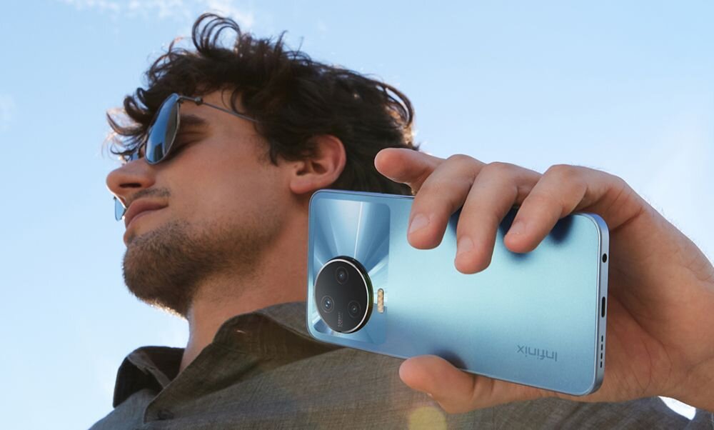 Osoba trzyma smartfon w kolorze niebieskim z widocznym logo \'Infinix\' i okrągłym modułem aparatu. W tle widać jasne niebo.