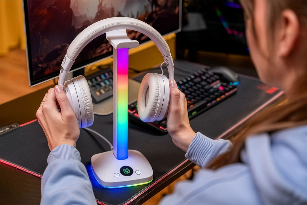 MAD DOG HPS800W Ujęcie z bliska, gdzie ręce trzymają białe słuchawki, które mają zostać umieszczone na stojaku MAD DOG. Stojak ma wielokolorowe podświetlenie RGB.