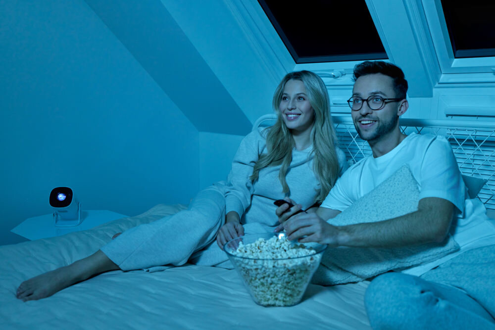 Projektor OVERMAX Multipic Veer - Wszechstronny projektor, ludzie leżący na łóżku, jedzący popcorn, oglądający wyświetlany obraz przez projektor