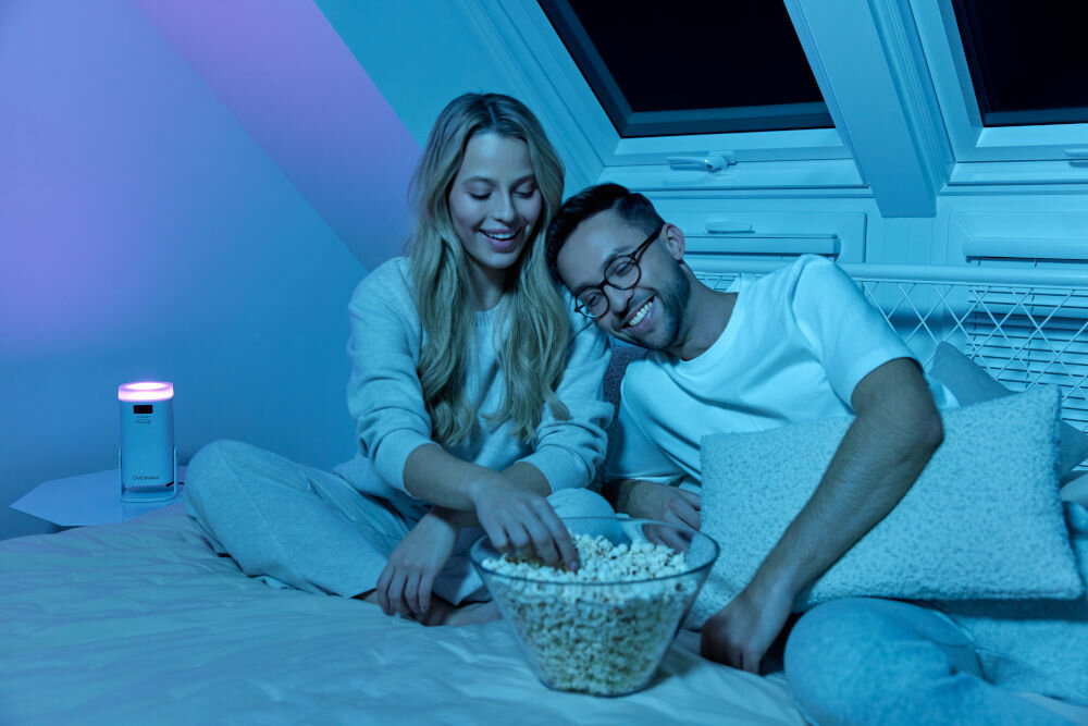 Projektor OVERMAX Multipic Veer - Łatwy w transporcie, projektor pod skosem, ludzie leżący na łóżku, jedzący popcorn, oglądający wyświetlany obraz przez projektor