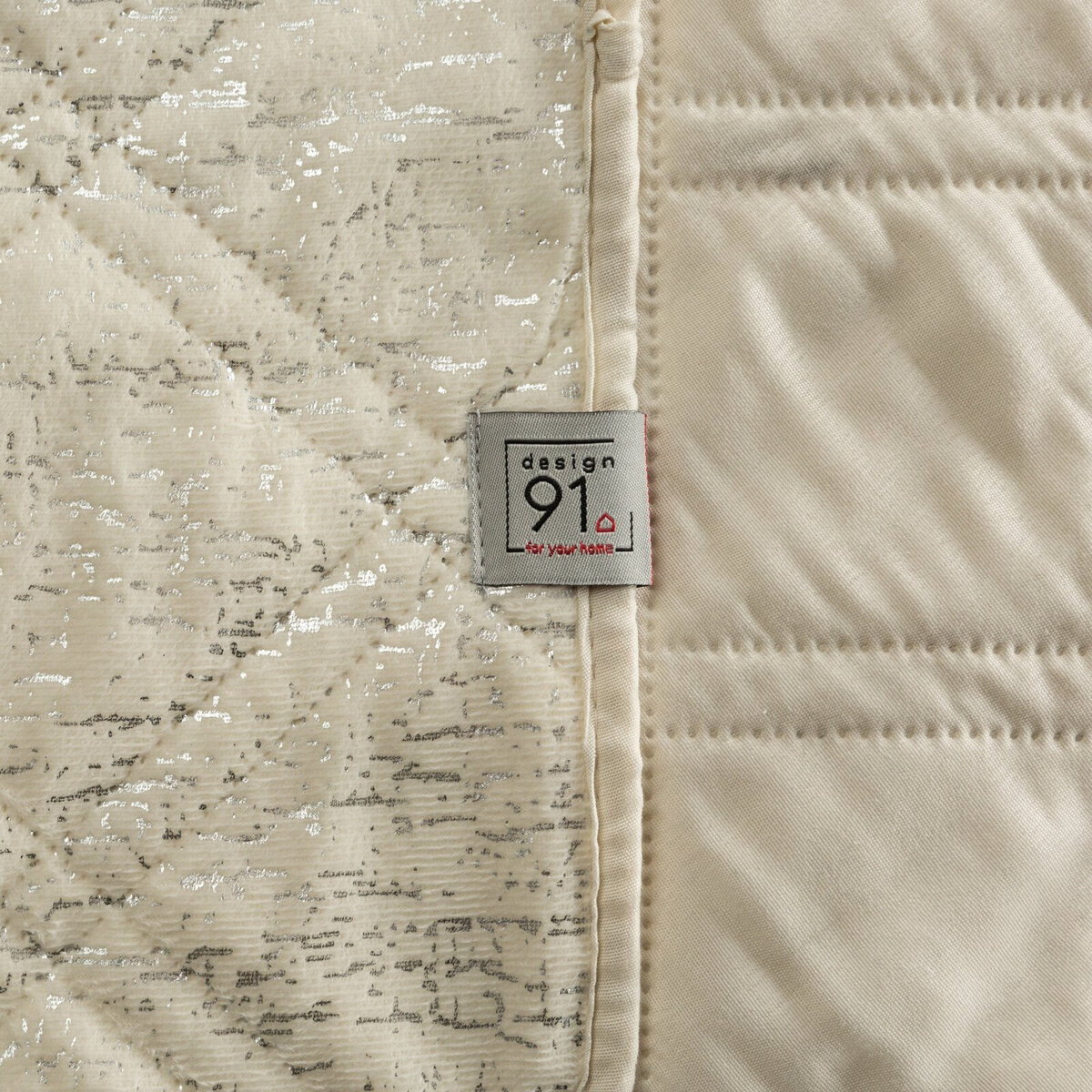 Obraz przedstawia fragment pikowanej narzuty w kolorze kremowym z połyskującymi elementami. Widoczna jest metka z napisem \'design 91 for your home\'.