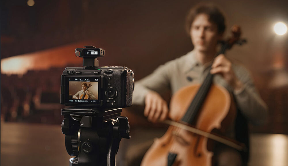Nadajnik DJI Mic 3 mężczyzna siedzący i grający na instrumencie strunowym na scenie przed nim kamera z podłączonym zestawem w tle sala widowiskowa automatyczne przełączanie częstotliwości z zasięgiem do 400 m i bezstratną transmisją audio jakość odporność na zakłócenia