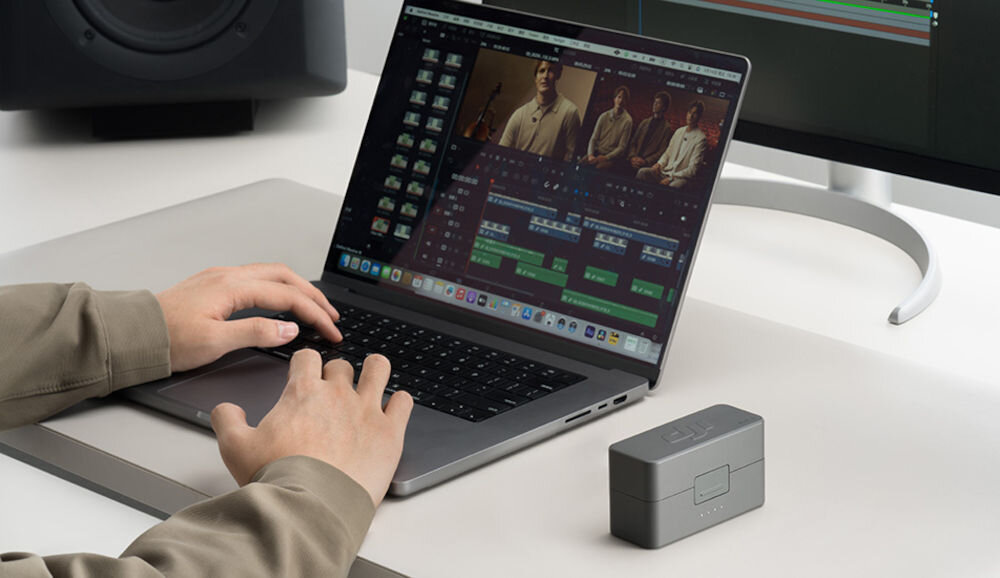 Nadajnik DJI Mic 3 prezentacja systemu w etui obok laptop i osoba siedząca przy montażu wideo w tle widoczny monitor i głośnik białe biurko system z funkcją timecode dokładnej synchronizacji nagrań z wielu kamer szybki i sprawny montaż DJI Mimo