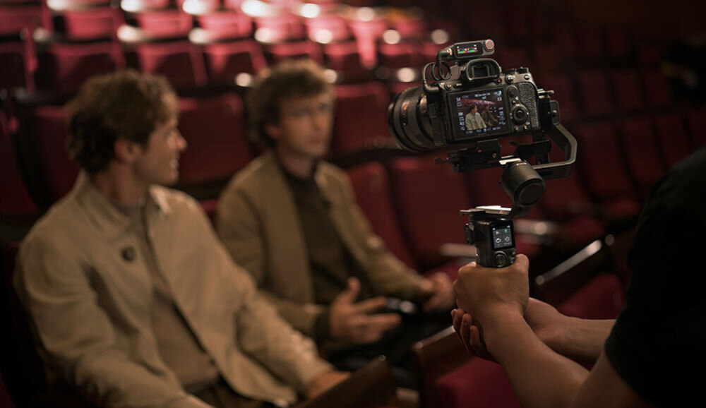 System bezprzewodowy DJI Mic 3 (2 TX + 1 RX + Etui ładujące) pokazanie zestawu podczas użytkowania podłączony do kamery i ubrań dwóch mężczyzn siedzących na fotelach w sali widowiskowej vlogi podcasty reportaże rozmowy grupowe mikrofon bezprzewodowy 4TX 8RX z 4 kanałami, adaptacyjnym wzmocnieniem i zapisem 32-bit float