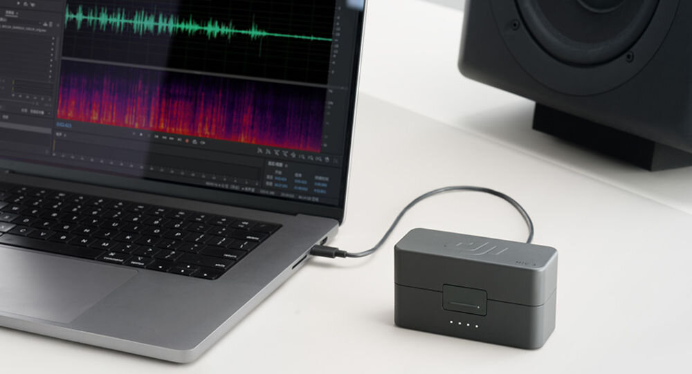Etui ładujące DJI Mic 3 prezentacja etui podłączonego do laptopa na ekranie laptopa program do edycji dźwięku w tle głośnik na białym biurku działanie przeznaczenie kompatybilność czas działania