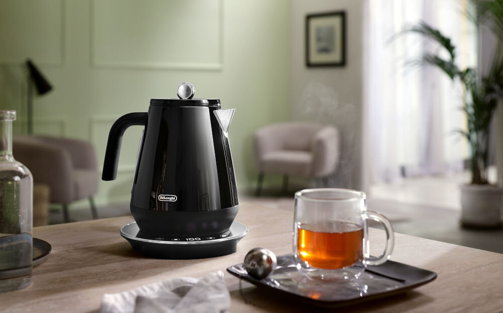 Czajnik DELONGHI KBY2011.BK czarny czajnik z podstawką stoi na blacie srebrne elementy obok parzy się herbata w przezroczystej szklance  