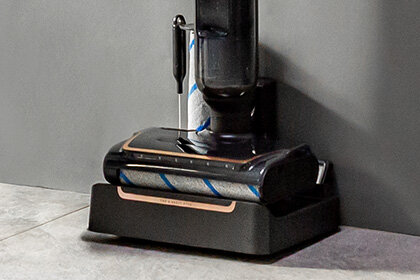 Bezprzewodowy odkurzacz X-CLEAN 7  w stacji czyszczącej ustawionej przy ścianie na jasnej, kafelkowej podłodze.