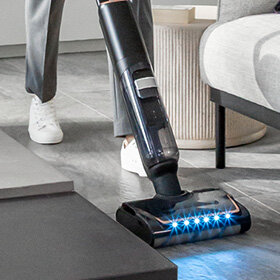 Bezprzewodowy odkurzacz X-CLEAN 7 z diodami LED czyści podłogę w salonie między sofą a stolikiem.