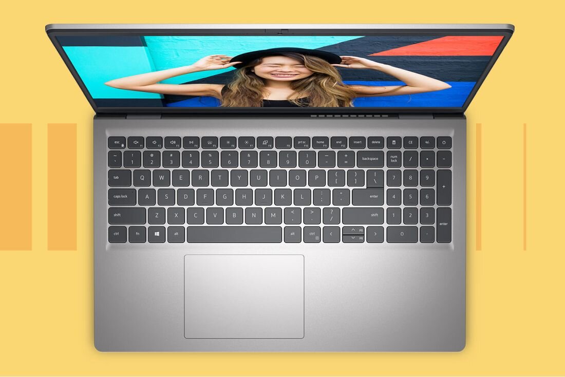 Laptop z otwartym ekranem, na którym widoczna jest osoba w kapeluszu na tle kolorowej ściany. Klawiatura jest pełnowymiarowa z wyraźnie zaznaczonym touchpadem.