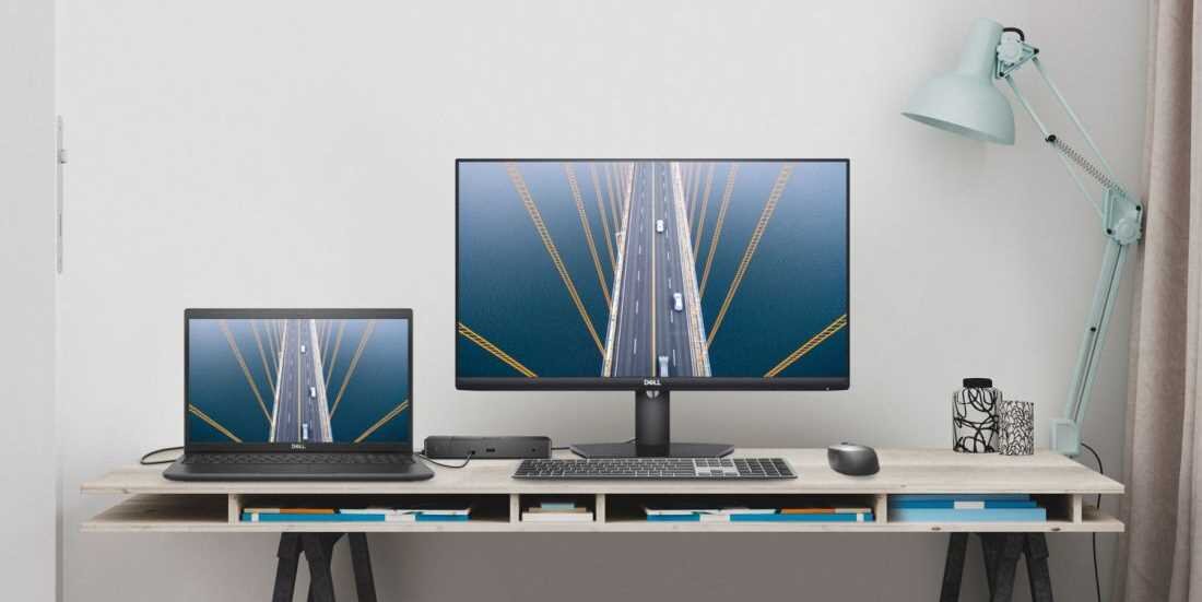 Na biurku znajduje się laptop oraz monitor, oba wyświetlają ten sam obraz przedstawiający most. Obok monitora stoi lampa biurkowa i kubek z długopisami.