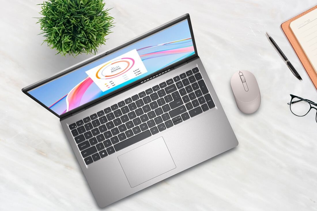 Na marmurowym blacie znajduje się otwarty laptop z widocznym ekranem, obok myszka, okulary, długopis i notes. Na ekranie laptopa widoczny jest kolorowy wykres z napisem \'Atlas\'.