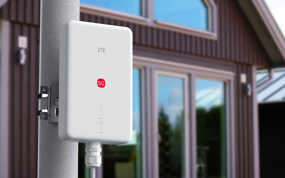 Router ZTE G51F 5G Biała jednostka zewnętrzna (ODU) routera ZTE G51F z czerwonym logo 5G jest zamontowana na słupku przy drewnianym domu z dużymi oknami. Urządzenie, podłączone płaskim kablem, symbolizuje instalację 5G FWA (Fixed Wireless Access), zapewniającą silny i stabilny dostęp do sieci w warunkach domowych.