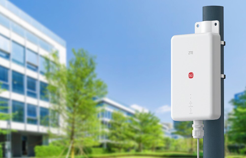 Router ZTE G51F 5G Router ZTE G51F zamontowany na zewnątrz budynku biurowego w otoczeniu zieleni. Ilustracja przedstawia urządzenie w zastosowaniu zewnętrznym – jako profesjonalne rozwiązanie dla firm, biur i sieci przemysłowych, zapewniające niezawodną łączność 5G.