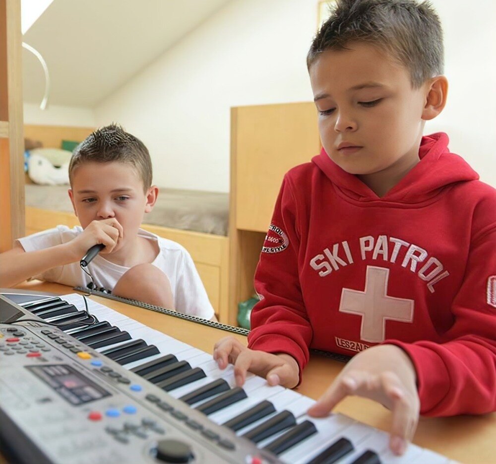 Dwóch chłopców bawi się przy elektronicznym keyboardzie; jeden z nich trzyma mikrofon, a drugi gra na klawiszach. Na bluzie jednego z chłopców widnieje napis \'SKI PATROL\'.