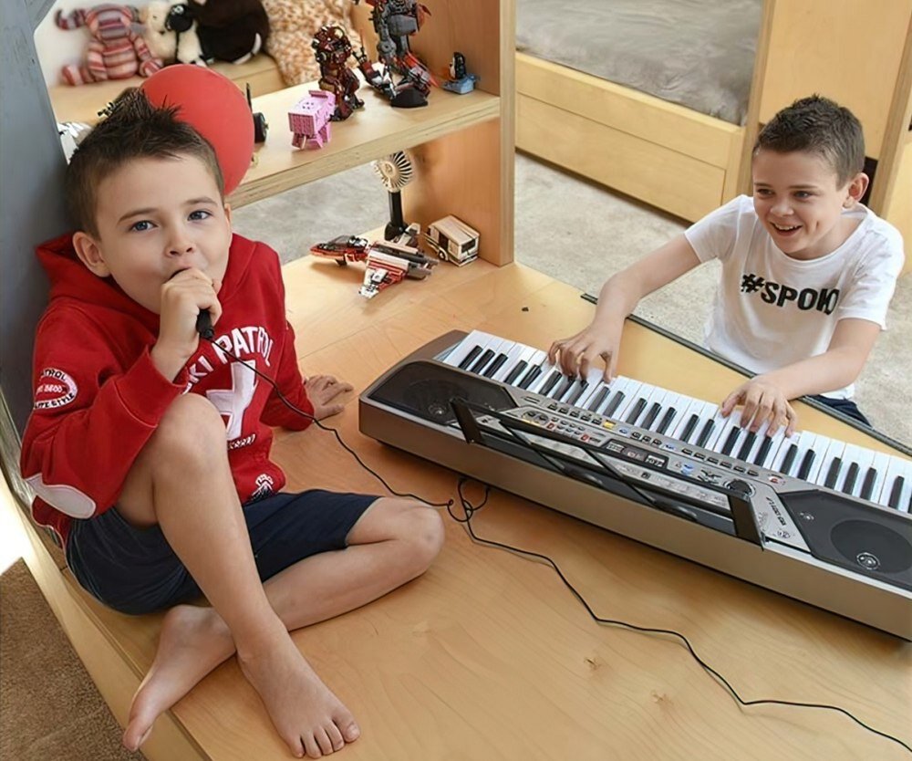 Dwóch chłopców bawi się przy elektronicznym keyboardzie, jeden z nich trzyma mikrofon. W tle widoczne są zabawki i półki z książkami.